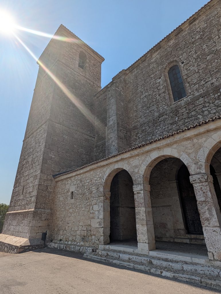 Picture of the sun-flared exterior of the Iglesia de San Pedro Apóstol in Los Santos de la Humosa, showing tower, portico, and side wall together in one frame.