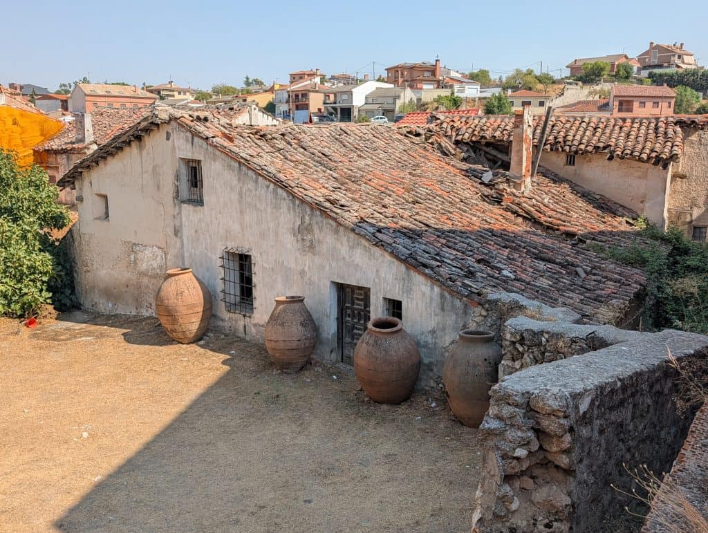 Picture of a Los Santos de la Humosa's old agricultural building with a partially collapsed tile roof and large ceramic storage jars in the foreground courtyard, modern rooftops visible beyond. 