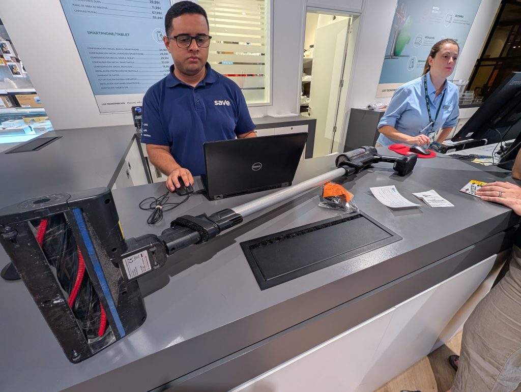 A Save-branded electronics service counter inside a Hipercor store. A technician in a navy polo shirt works at a laptop beside a disassembled cordless vacuum cleaner lying open on the counter, its base panel removed and internal wiring exposed. A price list for tech repair services is visible on the wall behind him, and a second staff member works at a nearby till.